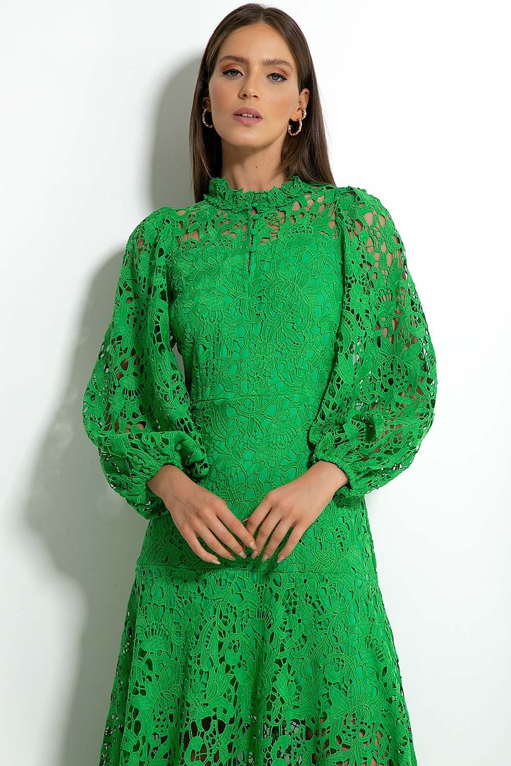 Green - A woman models a vibrant green, long-sleeve maxi dress featuring an intricate floral lace overlay, a high neckline, and a tiered skirt, holding a matching green clutch in a greenhouse setting.