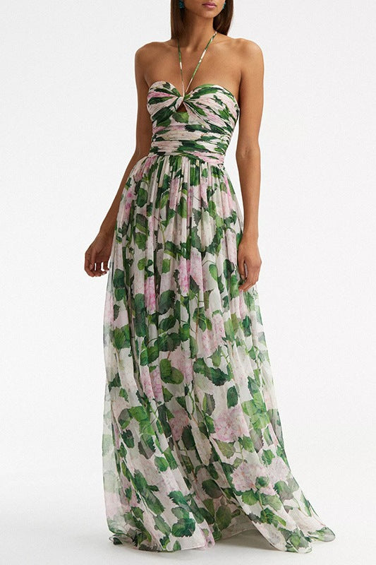 A model wearing a floor-length halter maxi dress in a white chiffon fabric featuring a green and pink hydrangea print, with a ruched bodice and flowing skirt.