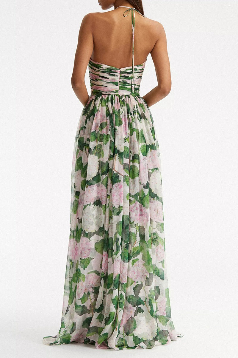 A model wearing a floor-length halter maxi dress in a white chiffon fabric featuring a green and pink hydrangea print, with a ruched bodice and flowing skirt.
