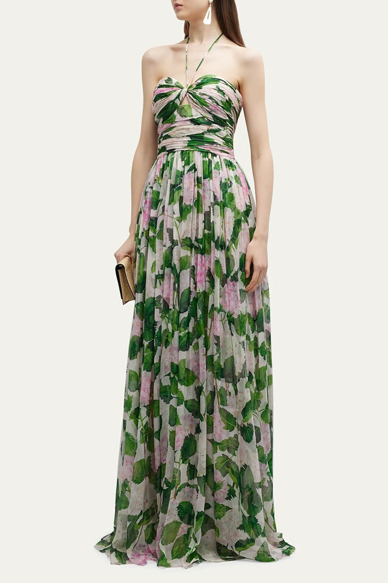 Green - A model wearing a floor-length halter maxi dress in a white chiffon fabric featuring a green and pink hydrangea print, with a ruched bodice and flowing skirt.