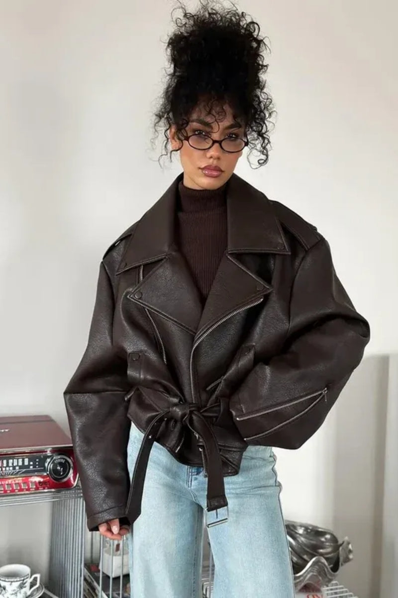 Brown - Woman wearing a dark brown oversized faux leather moto jacket with a tied waist belt, styled over a brown top and light wash jeans.