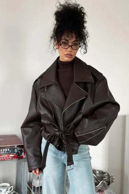 Brown - Woman wearing a dark brown oversized faux leather moto jacket with a tied waist belt, styled over a brown top and light wash jeans.