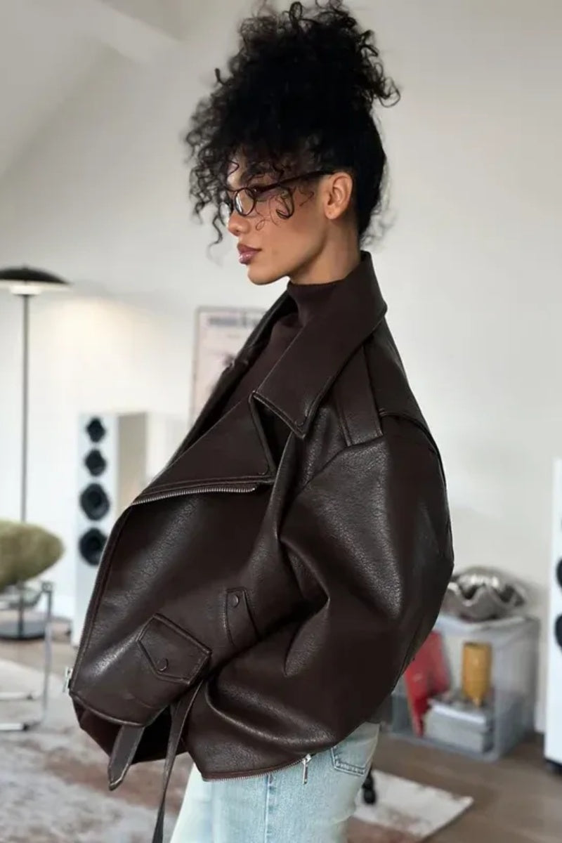 Brown - Woman wearing a dark brown oversized faux leather moto jacket with a tied waist belt, styled over a brown top and light wash jeans.