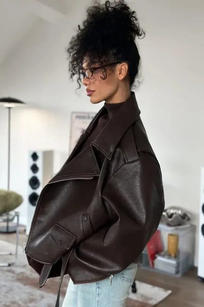 Brown - Woman wearing a dark brown oversized faux leather moto jacket with a tied waist belt, styled over a brown top and light wash jeans.