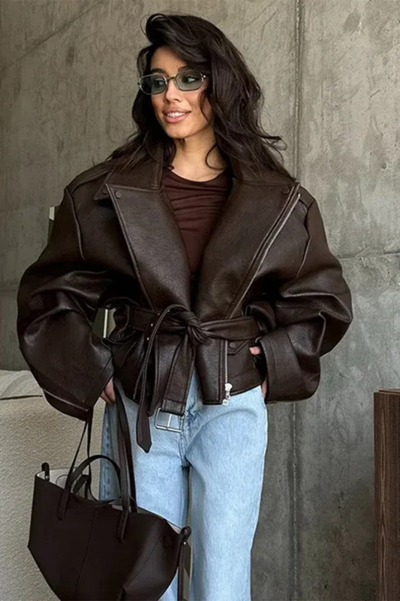 Brown - Woman wearing a dark brown oversized faux leather moto jacket with a tied waist belt, styled over a brown top and light wash jeans.