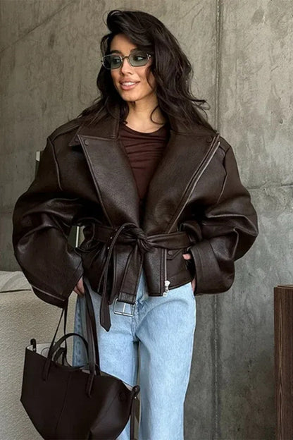Brown - Woman wearing a dark brown oversized faux leather moto jacket with a tied waist belt, styled over a brown top and light wash jeans.