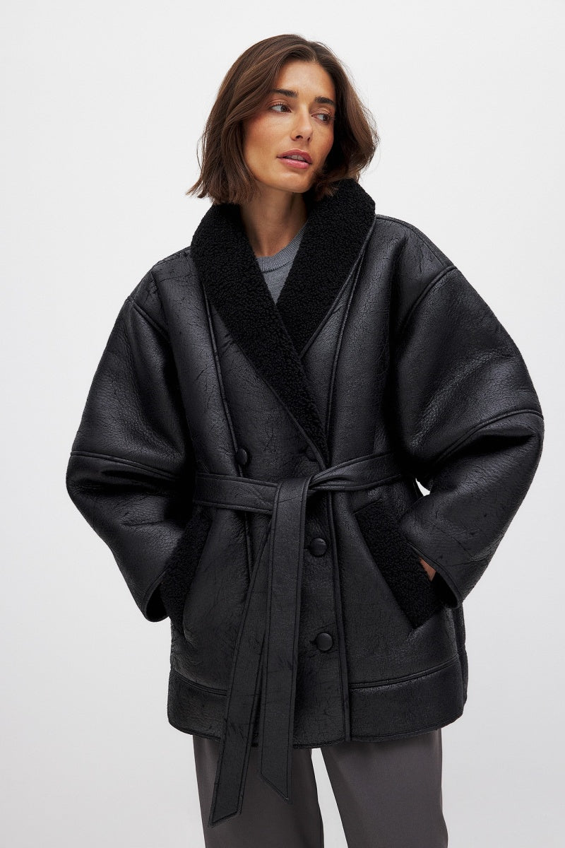 Black - A woman wearing a black, oversized faux leather coat with a plush faux shearling lapel, cinched at the waist with a self-tie belt, standing against a wood-paneled wall.