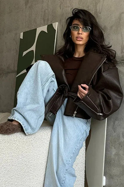 Brown - Woman wearing a dark brown oversized faux leather moto jacket with a tied waist belt, styled over a brown top and light wash jeans.