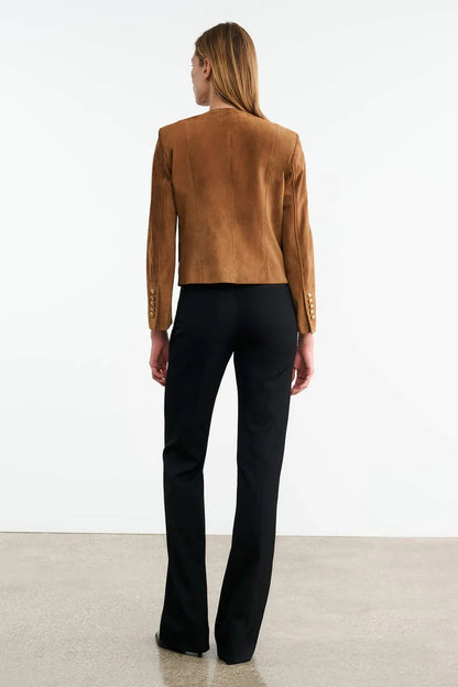 Brown - A woman wearing a tan, collarless suede cropped jacket with gold-tone buttons over a black top and trousers.
