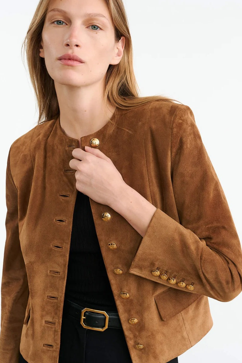 Brown - A woman wearing a tan, collarless suede cropped jacket with gold-tone buttons over a black top and trousers.