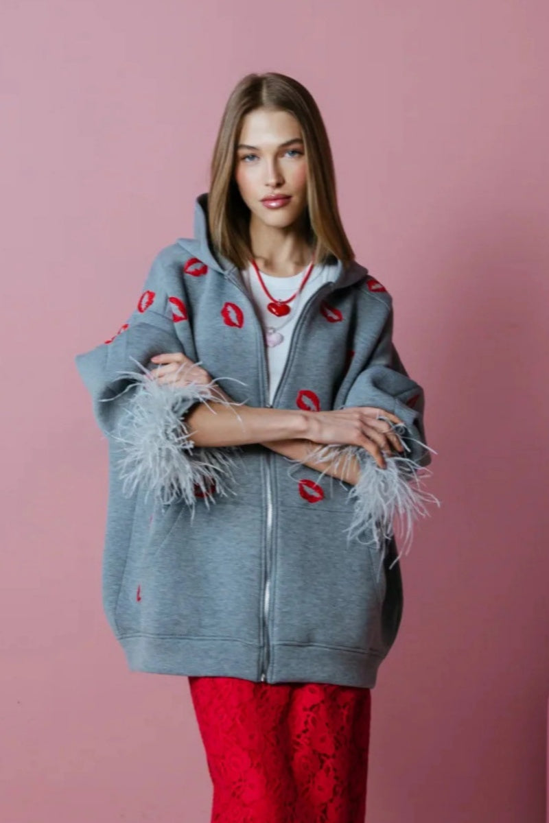Grey - A woman wearing an oversized grey zip-up hoodie featuring a red lip print and dramatic grey feather cuffs, styled over a white top and red textured skirt.