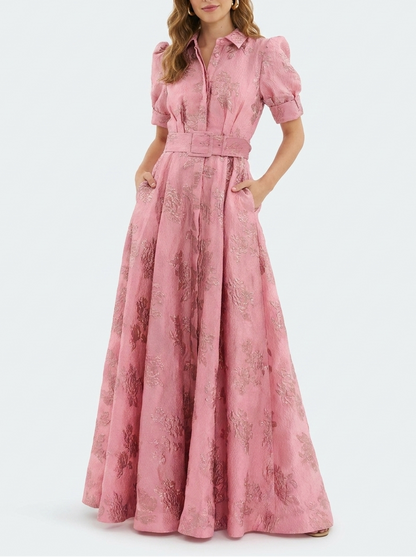 Woman wearing a long heavyweight jacquard maxi dress with a textured floral design, short puff sleeves, collared neckline, button-down front, and matching waist belt