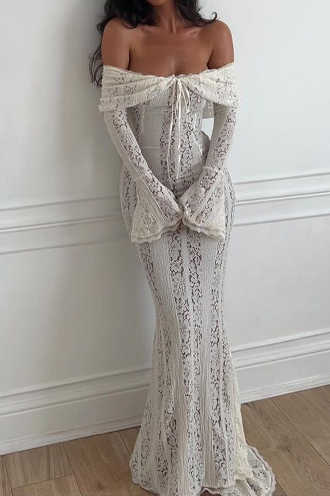 White - A woman wearing an off-white off-the-shoulder lace maxi dress with long flared sleeves and a tie-front detail, standing in front of a rustic wooden door.