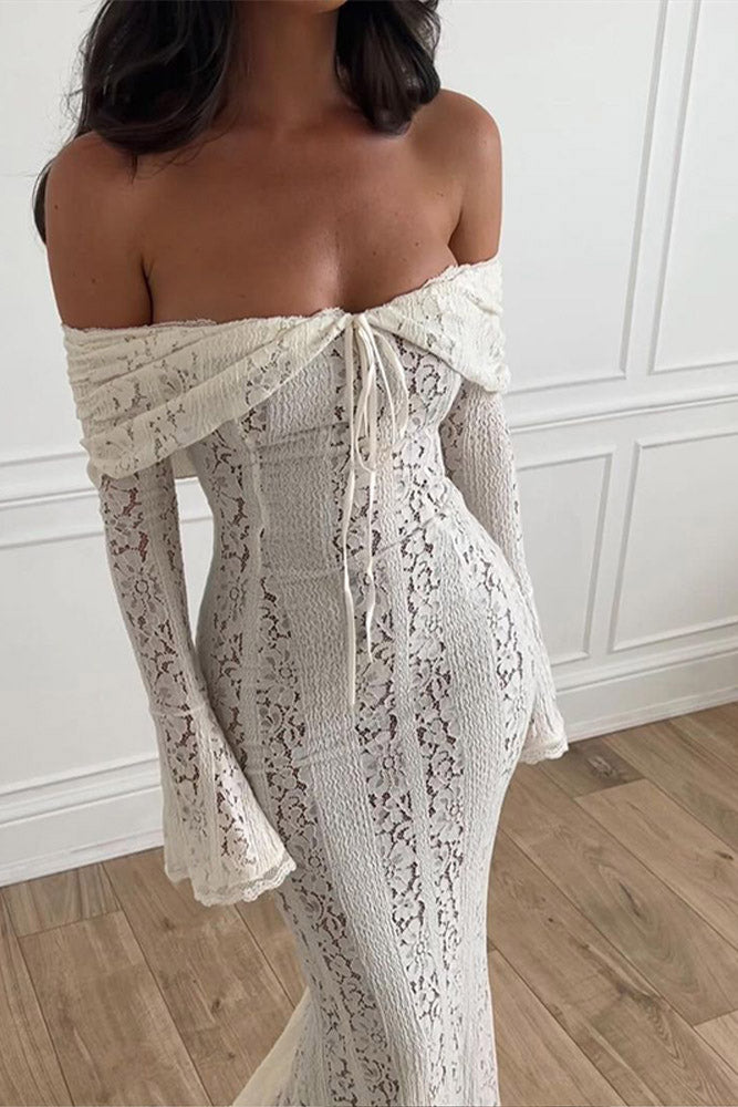 White - A woman wearing an off-white off-the-shoulder lace maxi dress with long flared sleeves and a tie-front detail, standing in front of a rustic wooden door.