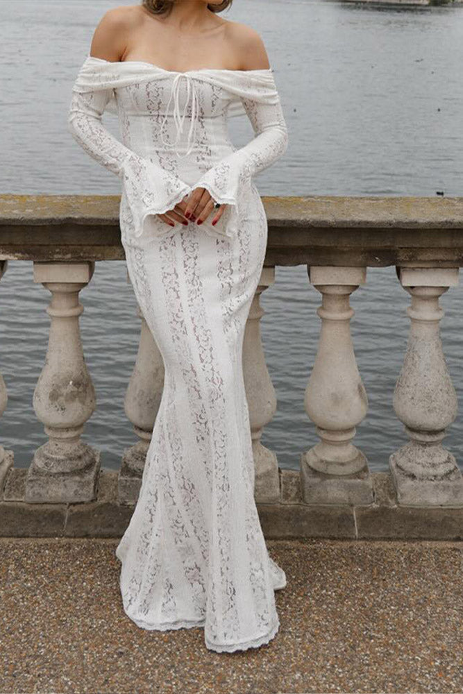 White - A woman wearing an off-white off-the-shoulder lace maxi dress with long flared sleeves and a tie-front detail, standing in front of a rustic wooden door.