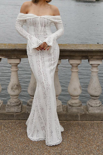 White - A woman wearing an off-white off-the-shoulder lace maxi dress with long flared sleeves and a tie-front detail, standing in front of a rustic wooden door.