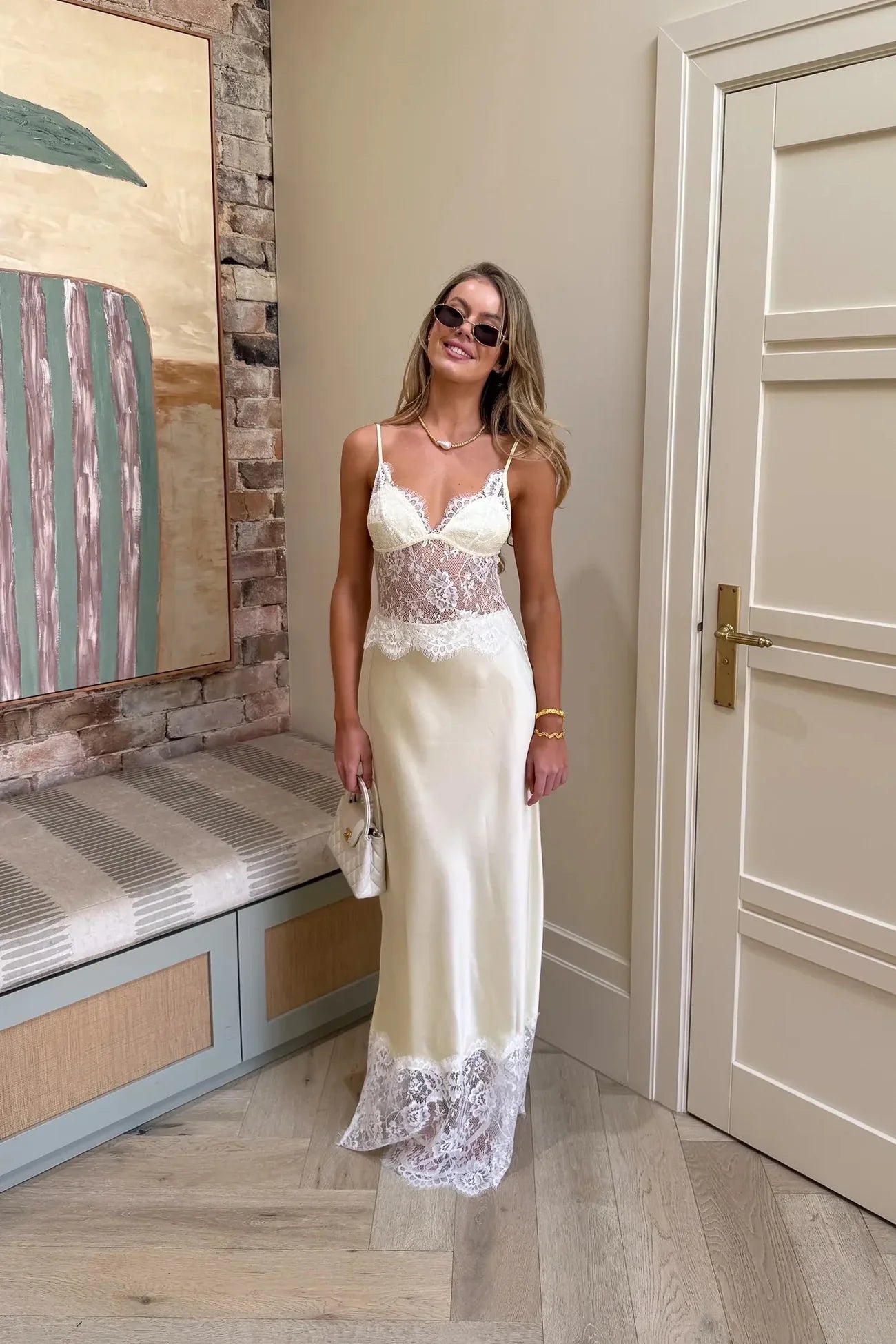A woman wearing a pale yellow maxi slip dress featuring a sheer floral lace bodice, spaghetti straps, and a lustrous satin skirt finished with a wide scalloped lace hem.