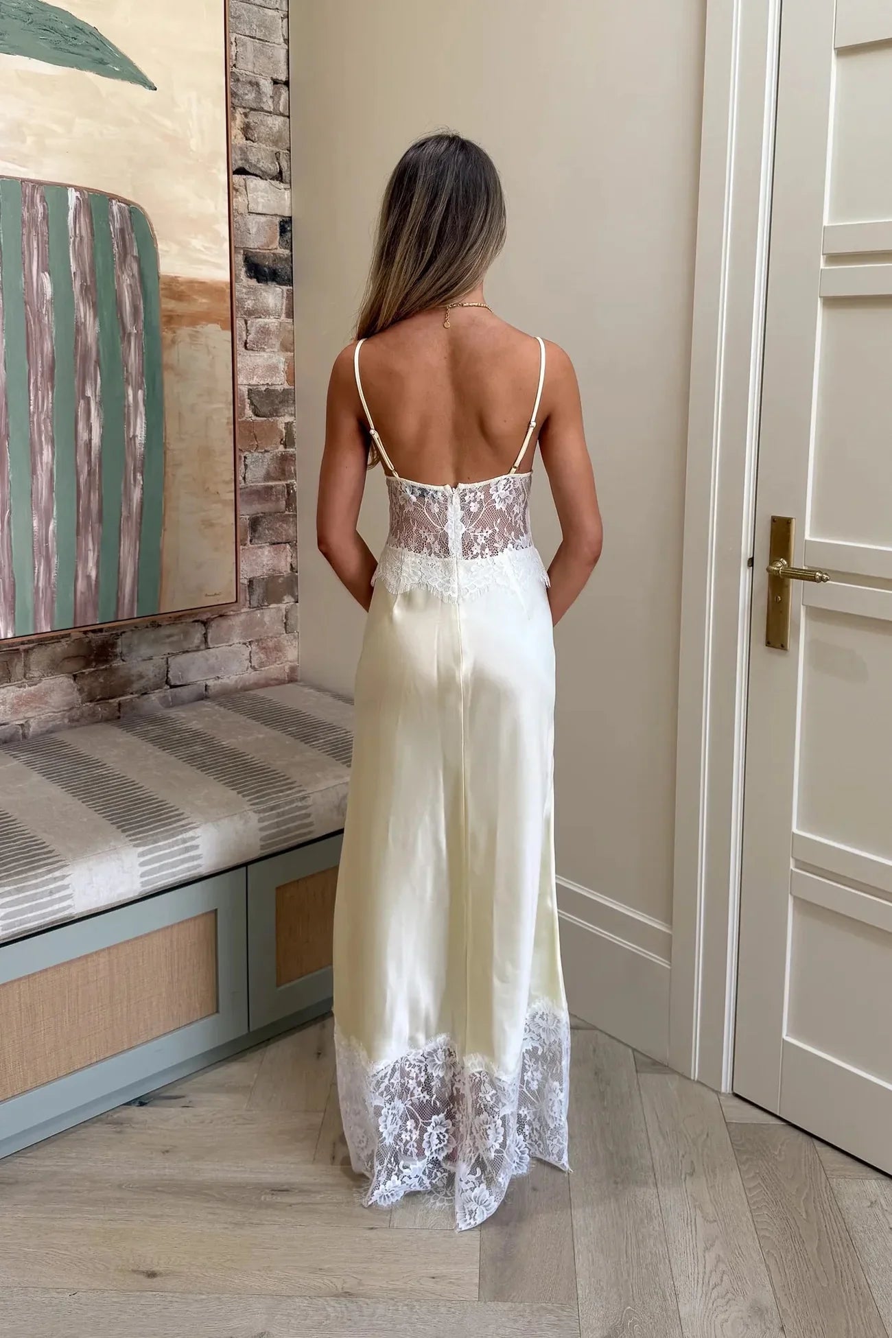 A woman wearing a pale yellow maxi slip dress featuring a sheer floral lace bodice, spaghetti straps, and a lustrous satin skirt finished with a wide scalloped lace hem.
