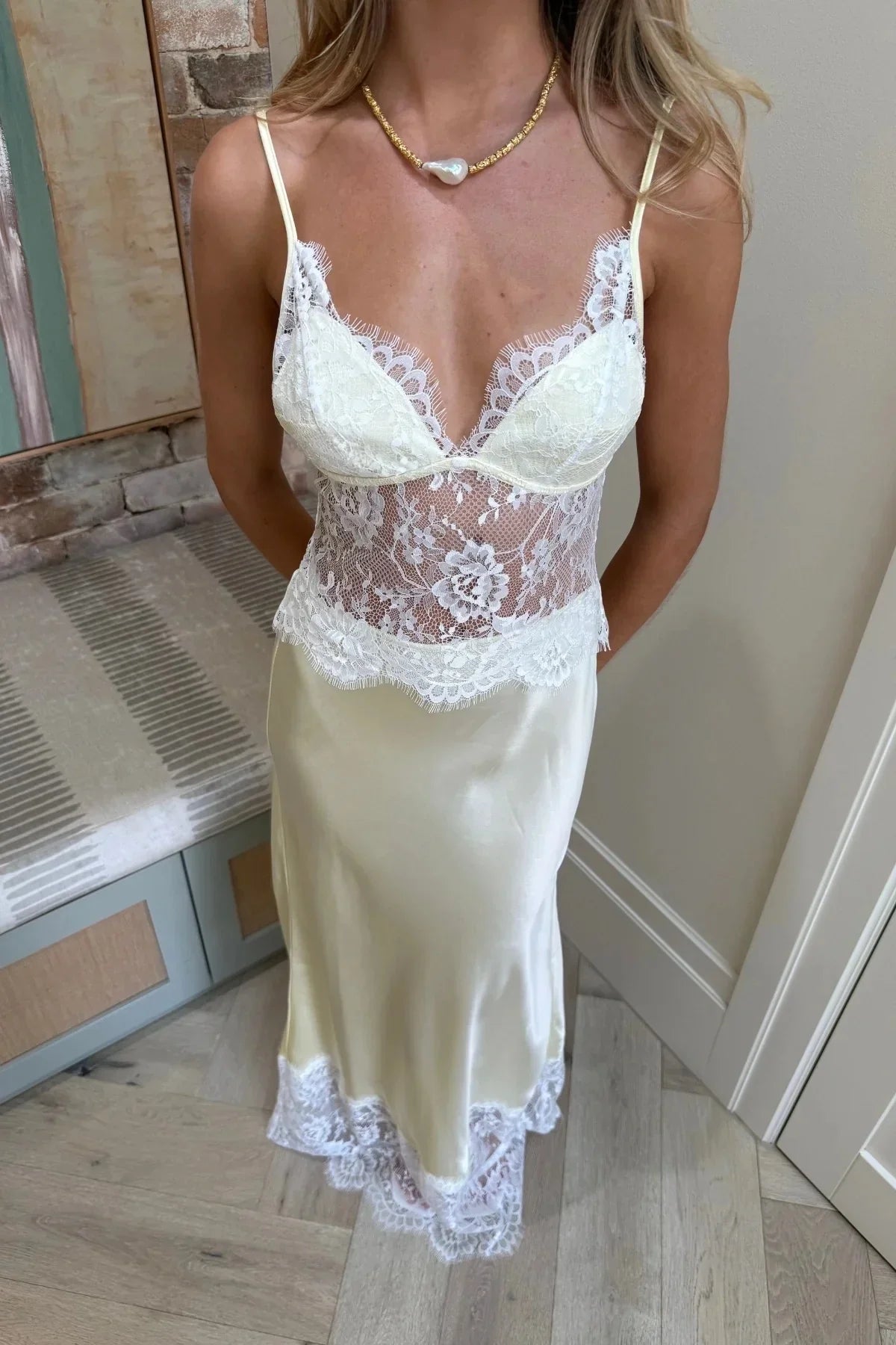 A woman wearing a pale yellow maxi slip dress featuring a sheer floral lace bodice, spaghetti straps, and a lustrous satin skirt finished with a wide scalloped lace hem.