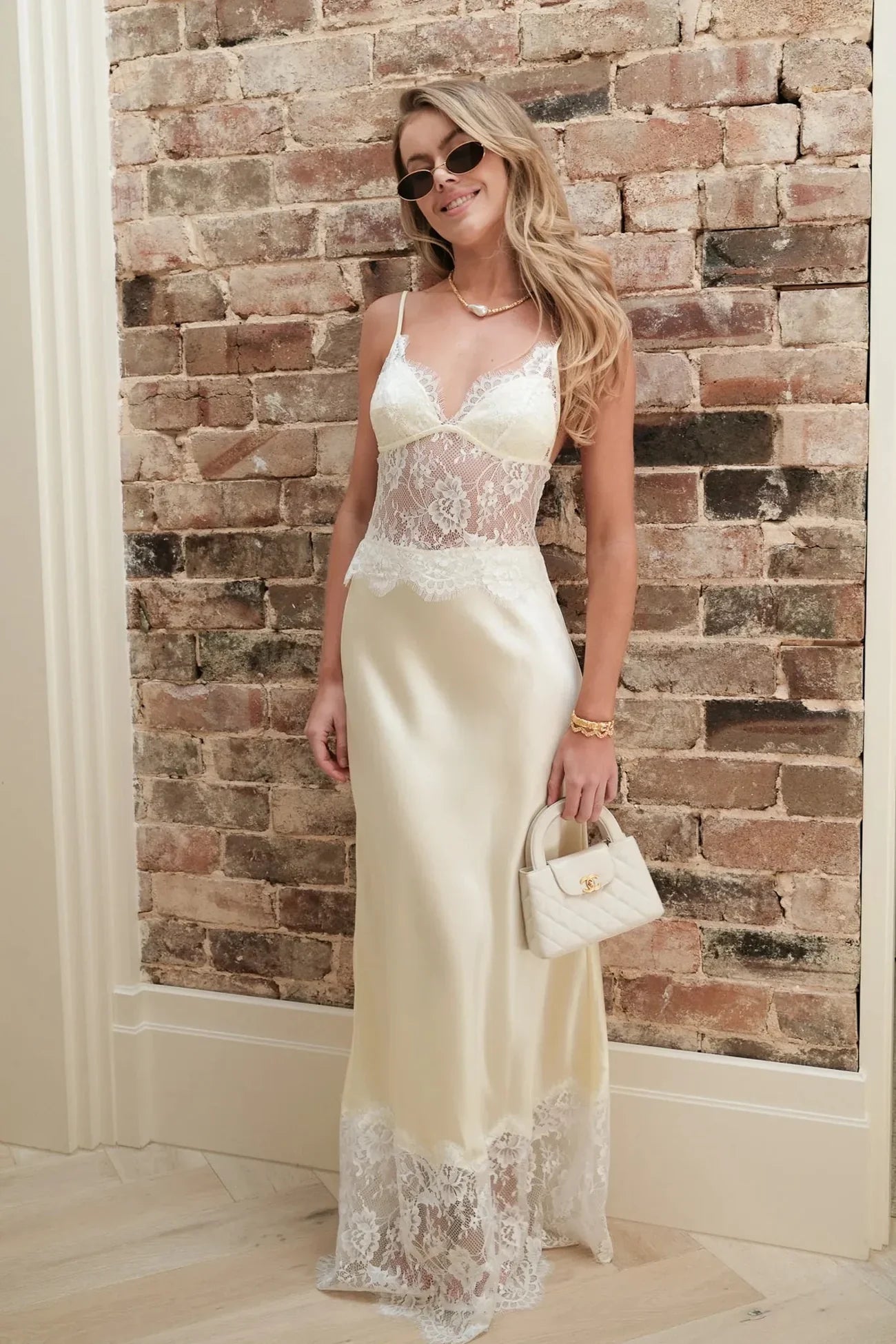 A woman wearing a pale yellow maxi slip dress featuring a sheer floral lace bodice, spaghetti straps, and a lustrous satin skirt finished with a wide scalloped lace hem.
