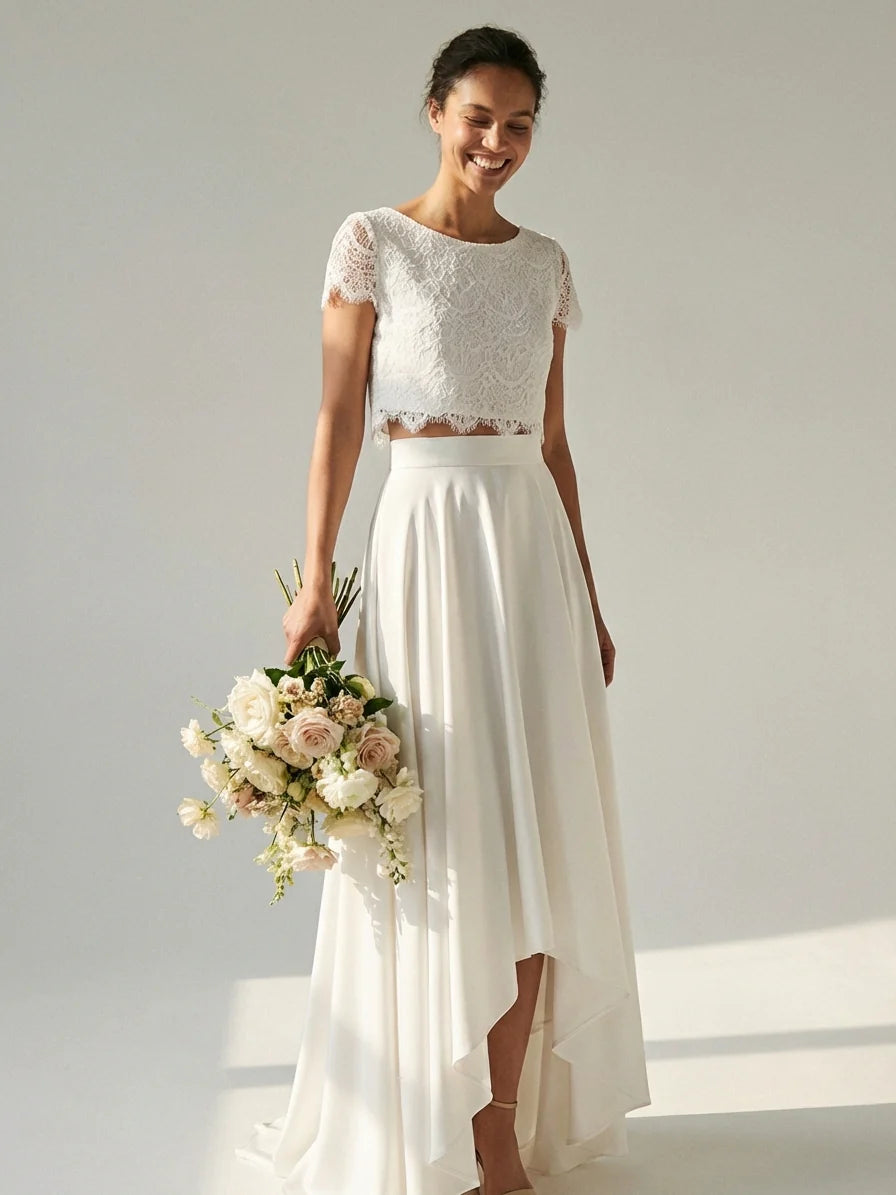 White - A smiling woman wearing a modern two-piece bridal set featuring a white short-sleeve lace crop top and a matching high-waisted, high-low white maxi skirt, holding a bouquet of white and blush roses.