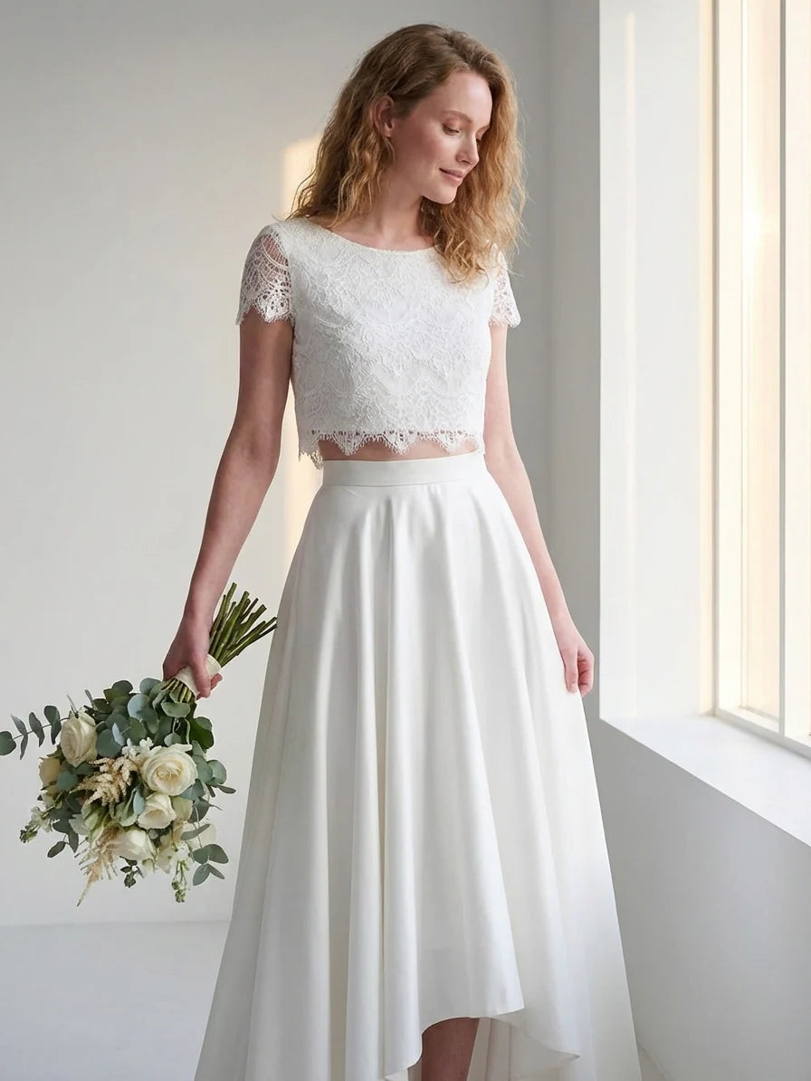 White - A smiling woman wearing a modern two-piece bridal set featuring a white short-sleeve lace crop top and a matching high-waisted, high-low white maxi skirt, holding a bouquet of white and blush roses.