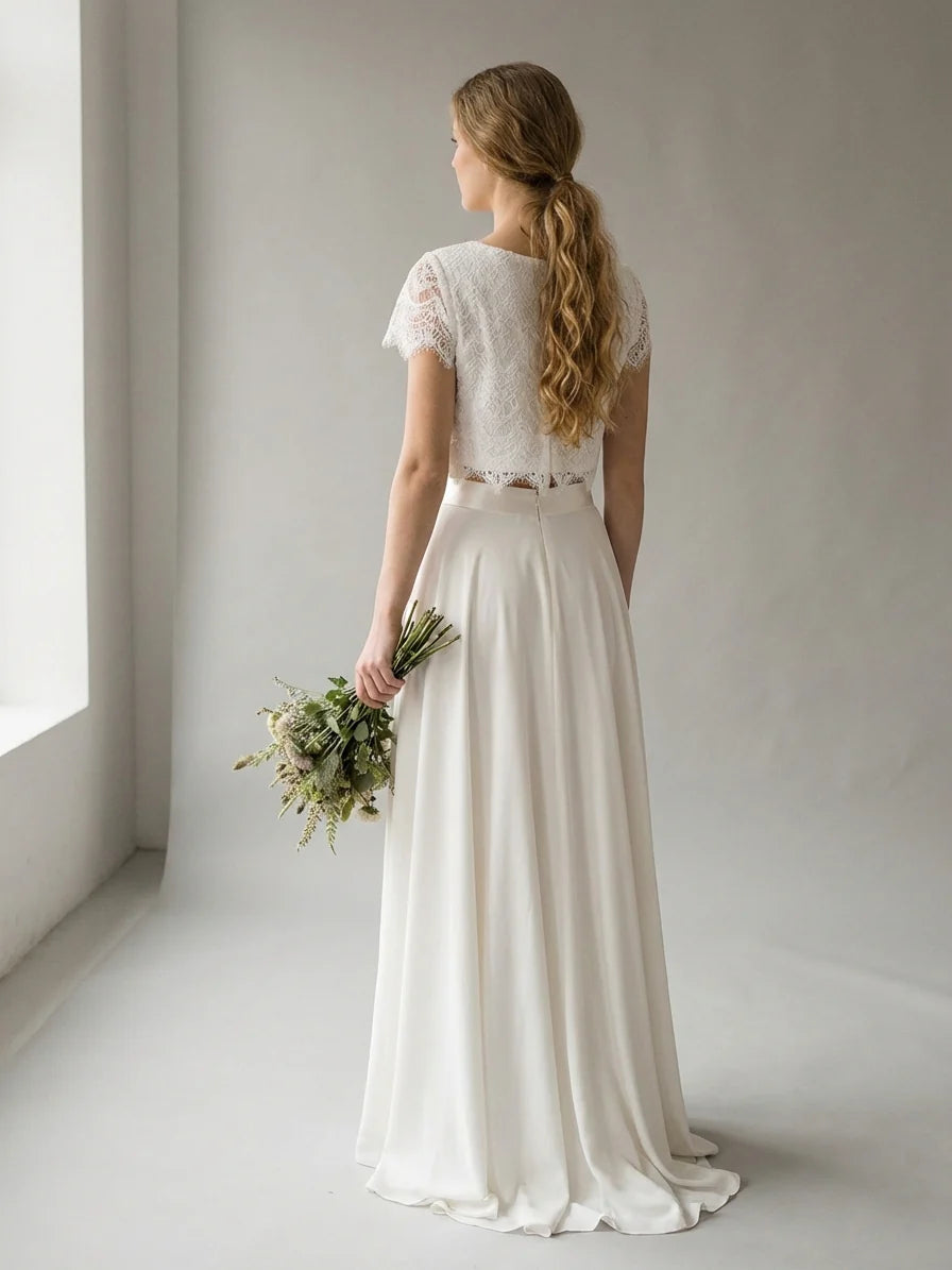 White - A smiling woman wearing a modern two-piece bridal set featuring a white short-sleeve lace crop top and a matching high-waisted, high-low white maxi skirt, holding a bouquet of white and blush roses.