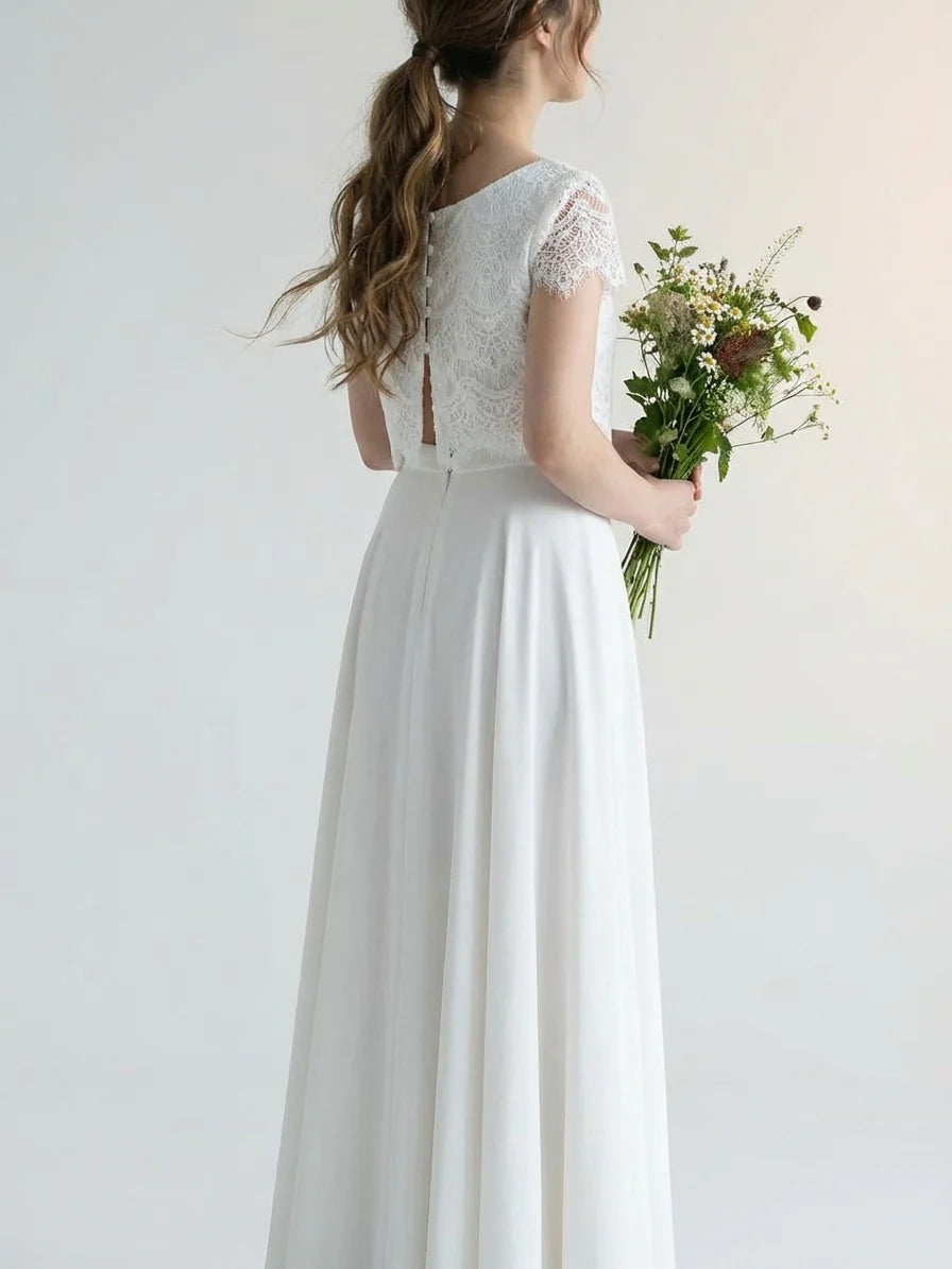 White - A smiling woman wearing a modern two-piece bridal set featuring a white short-sleeve lace crop top and a matching high-waisted, high-low white maxi skirt, holding a bouquet of white and blush roses.