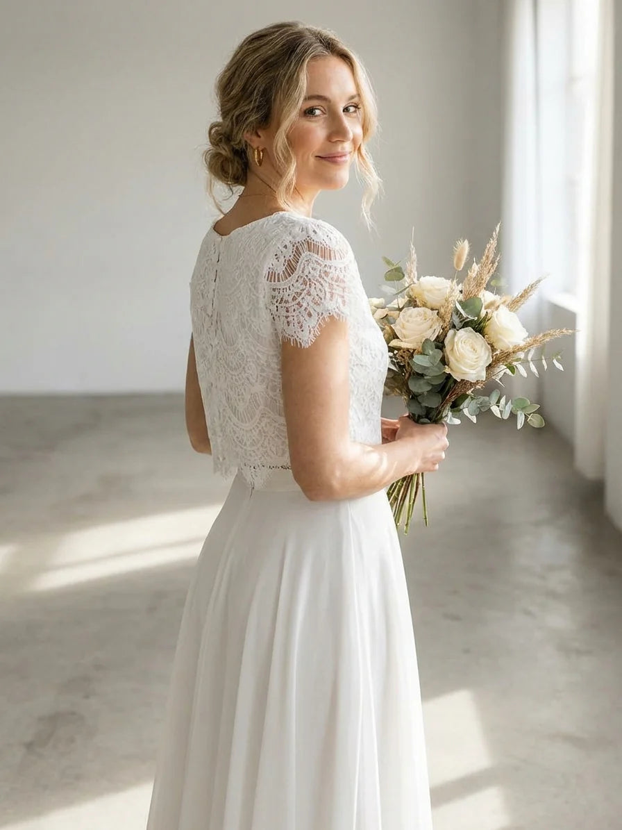 White - A smiling woman wearing a modern two-piece bridal set featuring a white short-sleeve lace crop top and a matching high-waisted, high-low white maxi skirt, holding a bouquet of white and blush roses.