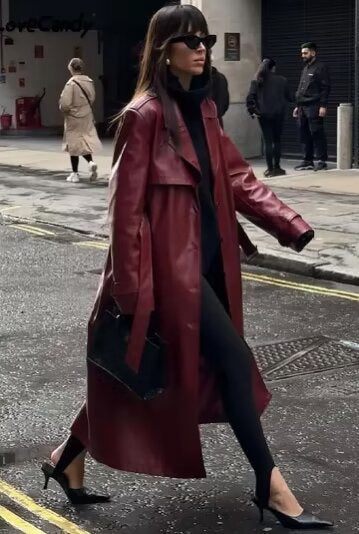 A woman walking down a city street wearing a deep burgundy maxi-length trench coat in a smooth leather-look material, styled with a black turtleneck, black stirrup pants, and black heels.