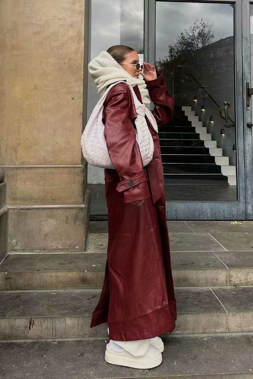 A woman walking down a city street wearing a deep burgundy maxi-length trench coat in a smooth leather-look material, styled with a black turtleneck, black stirrup pants, and black heels.