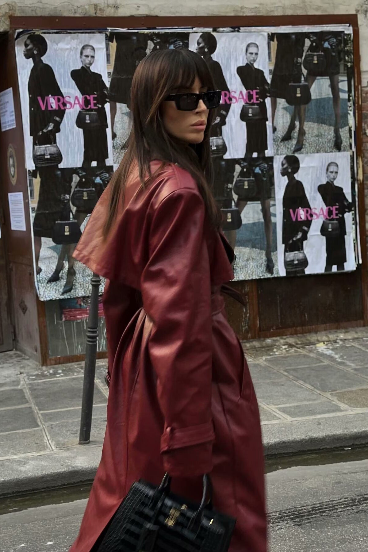 A woman walking down a city street wearing a deep burgundy maxi-length trench coat in a smooth leather-look material, styled with a black turtleneck, black stirrup pants, and black heels.