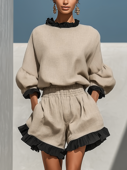 Beige - A model wearing an oatmeal-coloured linen two-piece set featuring a high-neck long-sleeve top and matching shorts, both detailed with contrast black ruffle trims along the collar, cuffs, and hems.