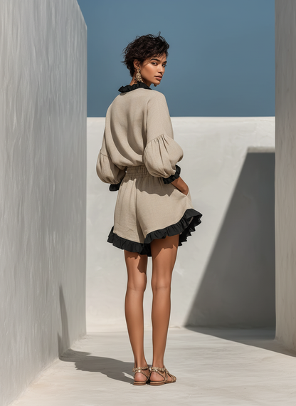 Beige - A model wearing an oatmeal-coloured linen two-piece set featuring a high-neck long-sleeve top and matching shorts, both detailed with contrast black ruffle trims along the collar, cuffs, and hems.