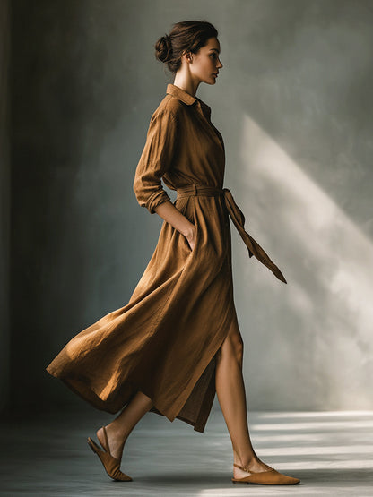 Brown - A woman walking in a long-sleeve, textured brown linen maxi shirt dress with a tied waist, button-up front, and side split, showcasing a fluid drape.