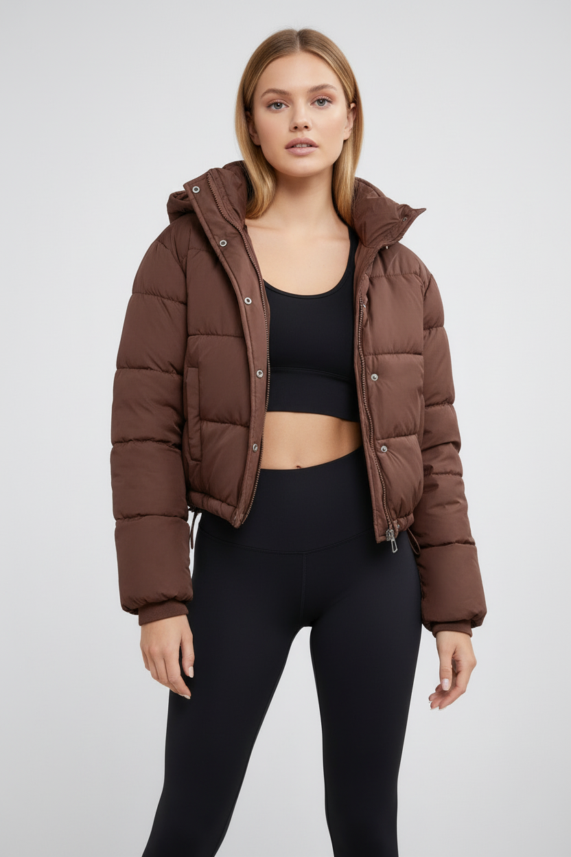 Brown - A woman wearing a chocolate brown cropped puffer jacket with a hood, styled over a black sports bra and high-waisted black leggings.