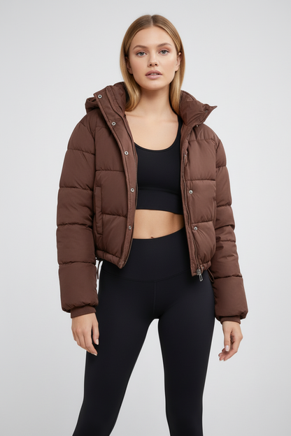 Brown - A woman wearing a chocolate brown cropped puffer jacket with a hood, styled over a black sports bra and high-waisted black leggings.