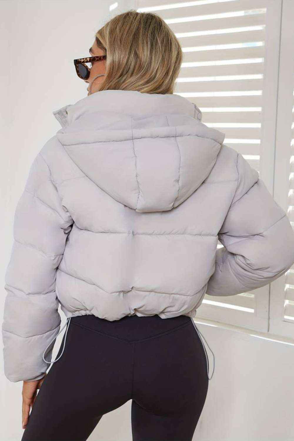 White - A woman wearing a chocolate brown cropped puffer jacket with a hood, styled over a black sports bra and high-waisted black leggings.