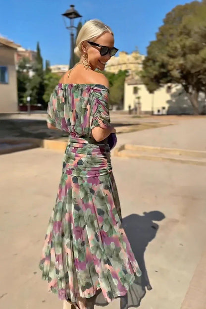 Multi - A woman walking outdoors wearing an elegant off-the-shoulder midi dress featuring a ruched bodice and a vibrant abstract floral print in shades of green, purple, and pink.