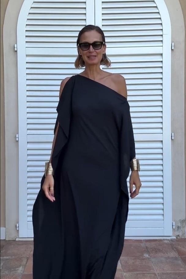A woman wearing a black one-shoulder asymmetric chiffon maxi dress with a flowing cape sleeve, styled with gold cuff bracelets and sunglasses against a white shuttered background.