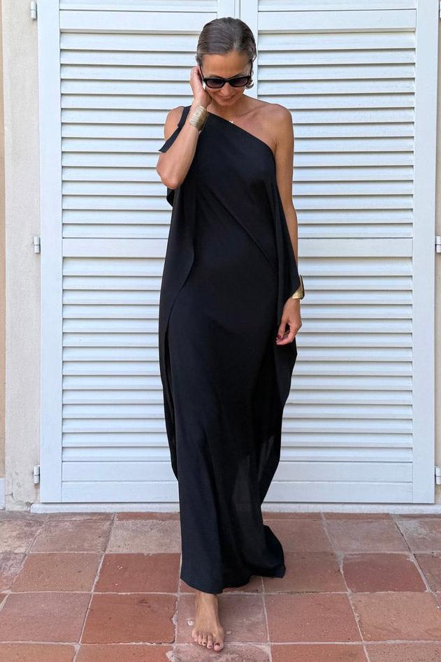 A woman wearing a black one-shoulder asymmetric chiffon maxi dress with a flowing cape sleeve, styled with gold cuff bracelets and sunglasses against a white shuttered background.