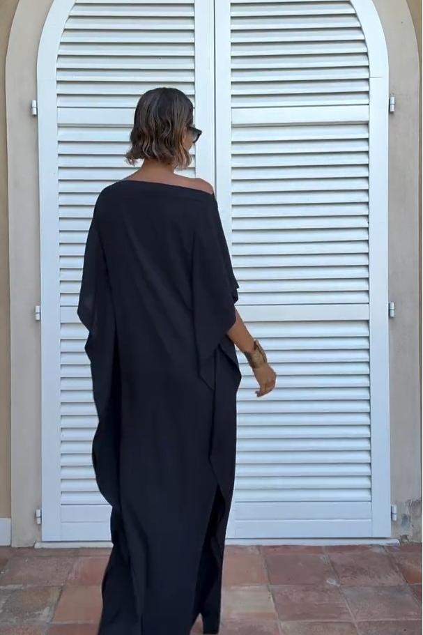 Black - A woman wearing a black one-shoulder asymmetric chiffon maxi dress with a flowing cape sleeve, styled with gold cuff bracelets and sunglasses against a white shuttered background.