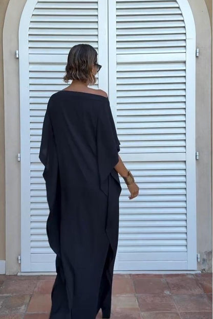 Black - A woman wearing a black one-shoulder asymmetric chiffon maxi dress with a flowing cape sleeve, styled with gold cuff bracelets and sunglasses against a white shuttered background.