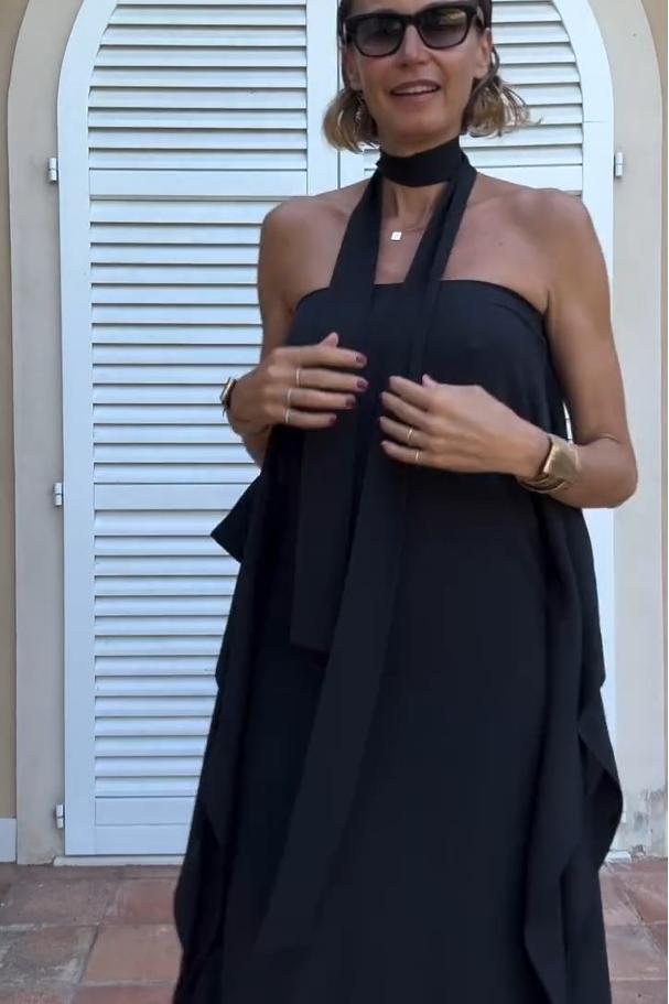 Black - A woman wearing a black one-shoulder asymmetric chiffon maxi dress with a flowing cape sleeve, styled with gold cuff bracelets and sunglasses against a white shuttered background.