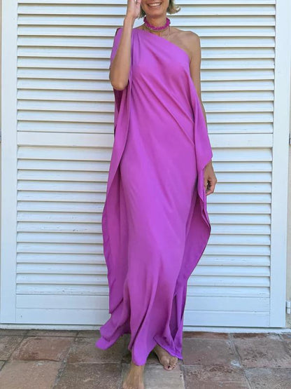 Cyclamen - A woman wearing a black one-shoulder asymmetric chiffon maxi dress with a flowing cape sleeve, styled with gold cuff bracelets and sunglasses against a white shuttered background.