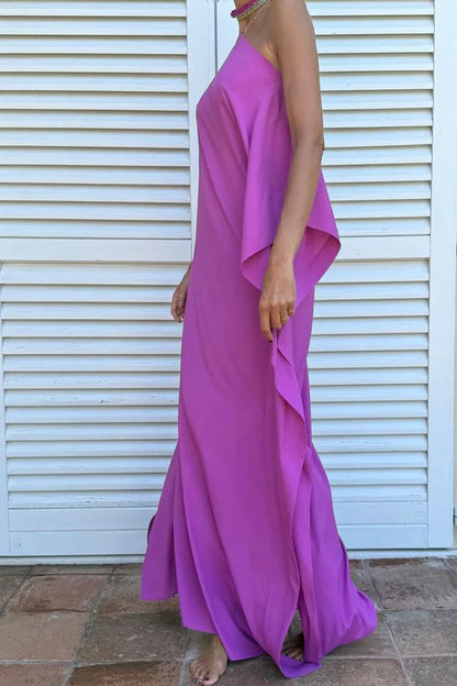 Cyclamen - A woman wearing a black one-shoulder asymmetric chiffon maxi dress with a flowing cape sleeve, styled with gold cuff bracelets and sunglasses against a white shuttered background.