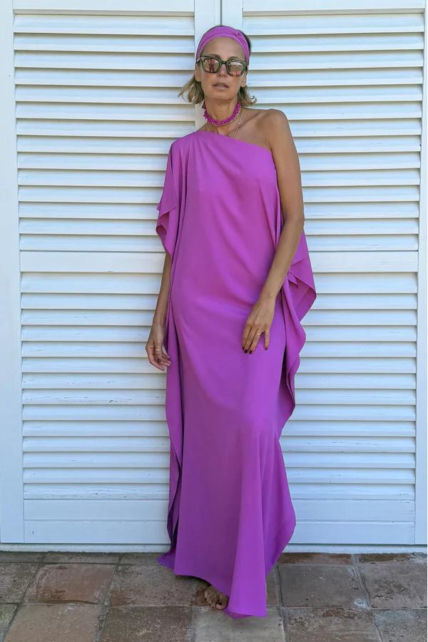 Cyclamen - A woman wearing a black one-shoulder asymmetric chiffon maxi dress with a flowing cape sleeve, styled with gold cuff bracelets and sunglasses against a white shuttered background.