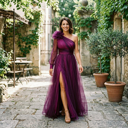 Dark Green - A woman wearing a floor-length, one-shoulder maxi dress in a green and purple abstract floral print, featuring a large 3D flower on the shoulder, a single sheer long sleeve, and a high leg split.