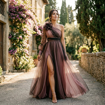 Pale Pink - A woman wearing a floor-length, one-shoulder maxi dress in a green and purple abstract floral print, featuring a large 3D flower on the shoulder, a single sheer long sleeve, and a high leg split.