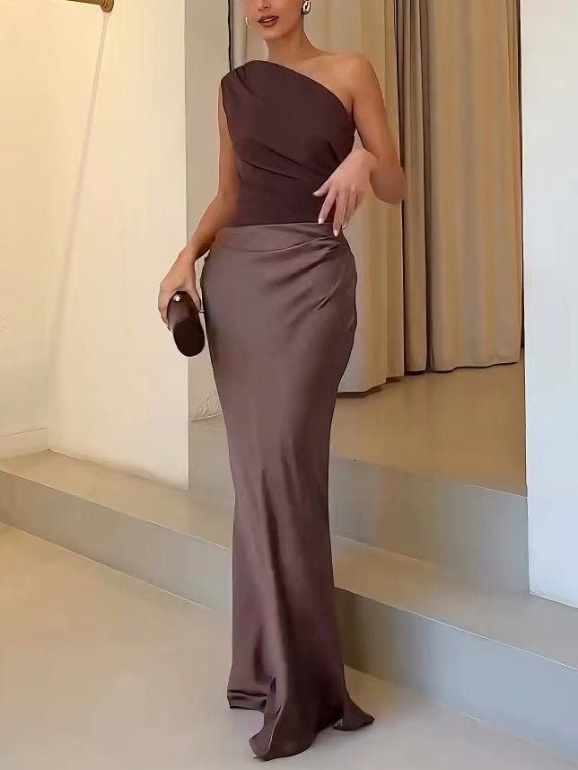 Blue - A woman wears a floor-length, one-shoulder maxi dress in a deep chocolate brown. The dress features a matte, draped bodice with an asymmetric waistline that flows into a lustrous, liquid-satin skirt.